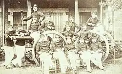 14 artilleurs en uniforme, assis devant, autour et sur un canon