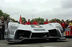 Photo d'une voiture vue de face, sur un sol asphalté. Une foule, des arbres et un ciel gris en arrière plan.