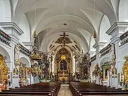 Une haute salle voûtée aux murs blancs et à la riche décoration dorée, avec des rangées de bancs de chaque côté d'une allée centrale.