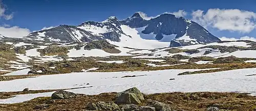 Paysage plat avec névés au premier plan et montagne en arrière-plan avec un glacier entouré de parois rocheuses verticales.