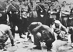 Photographie en noir et blanc de Juifs viennois, accroupis sur la chaussée, forcés d'effacer des slogans en faveur de la souveraineté de l'Autriche