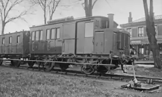 Photographie en noir et blanc de l'autorail de forme parallélépipèdique vue de trois quart face, côté droit visible, devant une gare. Il est accompagné de sa remorque. Des arbres sont présent le long du quai.