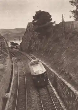 Photographie en sépia d'un autorail vu de dessus filant dans une tranchée percée d'un double voie et bordée de végétation méditerranéenne.