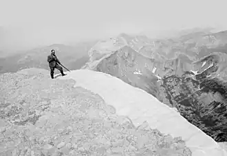 Photographie en noir et blanc d'un homme posant sur un sommet enneigé avec son bâton ferré.