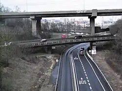 Ponts autoroutiers en béton et route à trois voies dont une voie de bus qui passe dessous, ville fortement urbanisée au loin.
