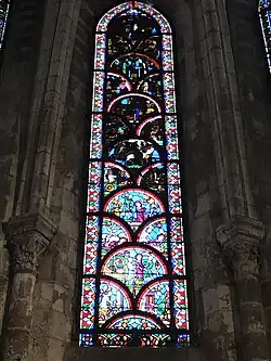 Vitrail composé de pièces arrondies en forme de coquillages. Sont représentés différents personnages dans plusieurs scénettes dont, en bas, un homme habillé de rouge s'agenouillant devant deux hommes levant le bras et semblant le menacer.  Les parties hautes sont difficilement visibles en raison du noircissement du vitrail.