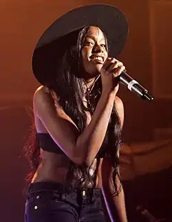 Une femme d'origine afro-américaine, Azealia Banks, tenant un microphone dans ses mains. Elle est vêtue d'une jeans bleu foncé de même que d'un soutien-gorge noir et porte un chapeau de la même couleur.