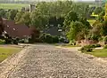 Photographie récente en couleurs d'une rue pavée, en pente, donnant en contrebas sur quelques habitations rurales.