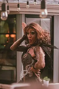 Photographie d'une drag queen noire en train de danser dans une terrasse de café. Elle porte une wig chocolat, une tenue noire et effectue des gestes et des expressions faciales maniérées.
