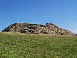 Vue de trois-quarts de l'ensemble du cairn, proue en premier plan. À la proue, la série de grosses dalles verticales du parement médian. Sur l'arrière du cairn, la partie détruite montre en coupe l'étagement des trois parements.