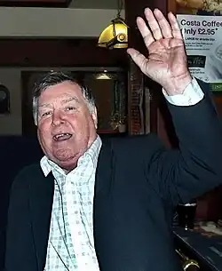 Portrait de face d'un homme debout dans un pub la main gauche levée, un fan le coude appuyé sur son épaule droite.