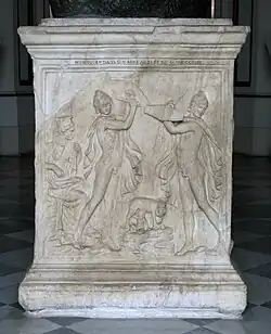 2 danseurs nus avec coiffe, cape, bouclier, épée, face à face, protègent un bébé assis au sol suçant le pis d'une chèvre