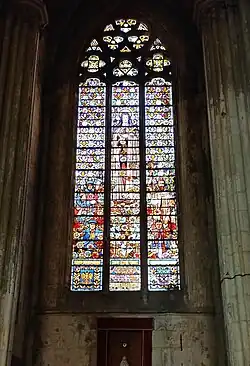Grand vitrail avec des teintes dominantes froides (bleu et vert surtout). Cinq personnages sont présents. Au centre de l'image, une femme avec une auréole porte une croix et lance des roses. En bas à gauche de l'image, un poilu de la Grande surmonte un marin en train de prier. À leur droite, un homme barbu tonsuré surmonte une femme à la peau mate en train de prier. Tout en bas, des écritures sont entourées de deux blasons.