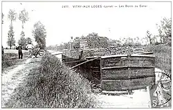 Carte postale noir et blanc du début du siècle. Un bateau, de type bâtard nivernais, une embarcation pouvant transporter 120&nbsp;tonnes de marchandises, est amarré au canal. Il est chargé de rondins de bois. Des ouvriers, à gauche, posent à côté de leur brouette spécifique pour le transport du bois, une brouette n’étant pas fermée sur le côté et pouvant transporter les rondins posés transversalement. Plusieurs planches posées transversalement entre le chemin de halage et le bateau font office de passerelle.