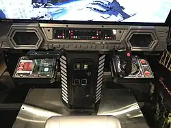 Sorte de meuble gris et noir imitant un cockpit de vaisseau, avec des boutons et des lumières, une manette de jeu de chaque côté et un monnayeur au centre.
