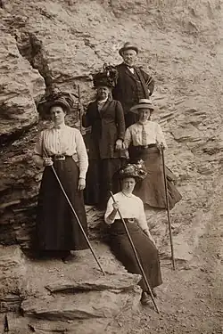 Quatre femmes en jupe longue et blouse blanche posent avec des bâtons de marche dans un paysage rocheux, accompagnées par un guide en arrière-plan.