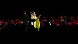 Image d'une chanteuse habillée en blanc et vert avec un masque florale jouant avec un orchestre