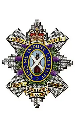 Image en couleur d'un insigne militaire composé d'une croix diamantée avec des listels d'or en haut et en bas ainsi qu'une couronne au naturel en haut. Au centre de l'insigne il y a un homme portant une robe brune tenant une croix blanche sur fond bleu entouré d'un anneau bleu foncé liséré d'or portant une inscription d'or entouré de chardon au naturel.