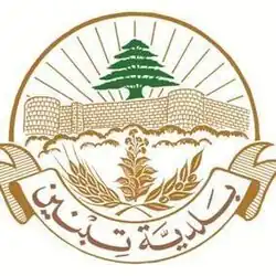 Description de Blason de Tibnine : Sur un fond blanc et en couleur vieux or, la partie supérieure des rayons de soleil en demi-cercle jaillissent de derrière le Toron des Chevaliers stylisé surplombé par un Cèdre vert du Liban. A la hauteur des fondations du château fort les sommets des arbres séparent le blason en deux. Une banderole ferme la partie inferieure portant l’inscription couleur bronze en arabe, municipalité de Tibnine. Dans son arrondi à dextre couché vers la droite, un rameau d'olivier en fruits, au centre une pousse de tabac à maturité, à sénestre couché vers la gauche, un épi du blé.