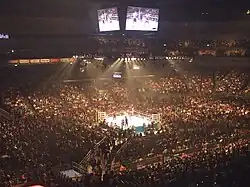 Deux combattants s'affrontent sur un ring éclairé au milieu d'une enceinte sportive dont les tribunes sont pleines.