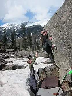 Un grimpeur sur un bloc en extérieur et une personne à côté du crash-pad qui fait une parade.