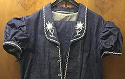 Photo d'une veste de bredzon pendue à un ceintre contre un mur en bois. La veste est de couleur bleue foncé avec des broderies d'edelweiss blanches.