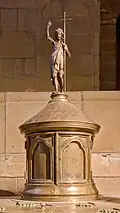 La statuette de Jean-Baptiste surmontant la cuve baptismale.