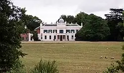 Photo d'un bâtiment à deux étages aux murs blancs et aux volets verts, entouré d'arbres, dans un parc à l'herbe rase où paissent quelques moutons