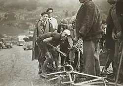 Photographie en noir et blanc montrant un cycliste regonflant son pneu, entouré par quelques hommes.