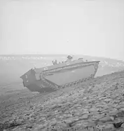 A tracked armoured vehicle drives up the bank of a river.  In the background lies the wrecked frame of a metal bridge.