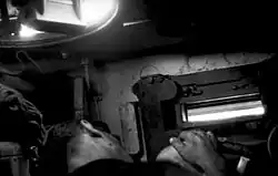 Photographie en noir et blanc montrant l’intérieur d’un poste de conduite avec un homme endormi à l’avant plan ; la seule vue sur l’extérieur est une étroite fente dans la paroi.