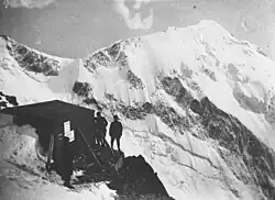 Photo ancienne d'un refuge entouré de trois silhouettes et une montagne enneigée en arrière-plan.