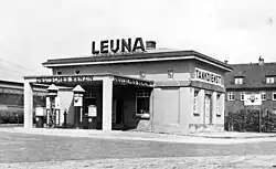 Photographie en noir et blanc montrant une station-service. Le mot « LEUNA » apparaît au-dessus de l'immeuble, alors que les expressions « Deutsches Benzin » et « Tankdienst » décorent certaines parties de l'immeuble.