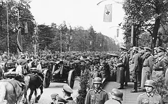 Photo noir et blanc prise le 5 octobre 1939, à Varsovie, en Pologne. Elle montre un défilé militaire de troupes de l'Armée allemande (à gauche) devant Adolf Hitler, debout et bras droit tendu devant lui, avec, dans son dos, une rangée d'officiers (à droite). Des arbres et un ciel clair forment l'arrière-plan de la photo.
