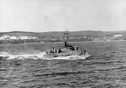 Navire avançant dans une mer agitée et laissant une large vague derrière lui. Des falaises et des collines boisées sont visibles à l'arrière-plan.