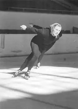 Helga Haase, portant un fichu sur la tête, pendant une course en intérieur. On voit le bras et le patin d'une concurrente derrière elle.