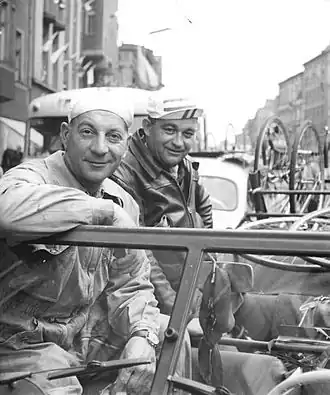 Photo noir et blanc de deux hommes accoudés sur le pare-brise d'une automobile suivant une course cycliste.