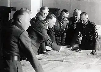Photo noir et blanc. Dans une pièce aux murs clairs, sept officiers généraux en uniforme suivent les indications données par Adolf Hitler pointant de sa main droite un lieu sur une carte étalée devant eux sur une table. Les huit hommes sont debout autour de la table ; Adolf Hitler et le général Friedrich Paulus sont au centre de la photo.