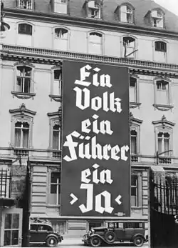 Photo noir et blanc, prise à Berlin en novembre 1933, d'une affiche électorale du NSDAP déployée sur les trois premiers étages de la façade d'un bâtiment de cinq étages (bureau du parti nazi). On peut lire, en grosses lettres, le slogan en allemand : « Ein Volk, ein Führer, ein Ja ».