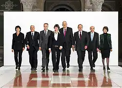 Photographie d'un groupe de cinq hommes et trois femmes marchant vers la caméra sur un fond blanc et sur un sol représentant le drapeau suisse.
