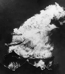 Photo noir et blanc d'un donjon de château en flammes, vu de nuit. Une colonne de feu s'élève dans le ciel sombre, du centre de l'image vers la droite.