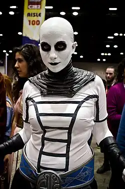 Femme portant un maquillage blanc intégral, à l’exception de rond noirs autour des yeux. Elle est vêtue de noir.