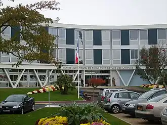 Photographie du CAREL, montrant un bâtiment parallélépipédique horizontal, légèrement concave, desservi par une volée de marches encadrées de massifs. Le rez-de-chaussée vitré est soutenu par des grands V de béton. Les deux étages sont faits d'une alternance de baies vitrées d'aluminium et de panneaux rectangulaires foncés. Les différents niveaux sont soulignés de larges bandeaux gris. Au-dessus de l'entrée principale est écrit en larges lettres capitales « Centre audiovisuel de Royan pour l'étude des langues ».