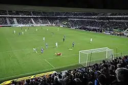 Vue générale du terrain depuis le derrière d'un but, l'action se déroulant sur le but du premier plan.