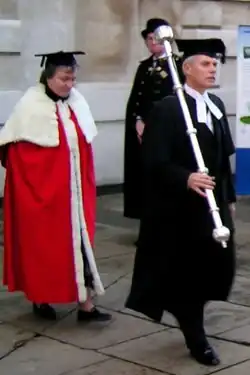 Universitaire portant la robe rouge et le mortar board ou square, sans doute l'invitée d'honneur, précédée du bedell en toge noire et square, avec dans les mains le mace