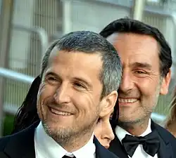 Deux hommes en smoking, souriants, l'un à moitié caché derrière l'autre.