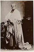 Photographie en noir et blanc du cardinal, de pied, en tenue ecclésiastique.