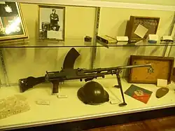 Photo en couleurs d'une mitrailleuse et d'un casque posée sur une tablette ; au-dessus, posés sur une autre tablette, il y a d'autres objets incluant un cadre avec un portrait et un autre cadre avec des médailles.