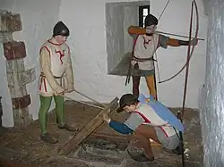 Photographie de plusieurs mannequins en habits médiévaux dans une salle aux murs blancs. L'un tire à l'arc par une meurtrière et les deux autres soulèvent une trappe en bois sur le sol.