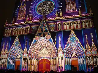 Vue en couleurs d’une façade de cathédrale, prise de nuit et illuminée de multiples couleurs.